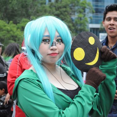 Person in blue wig and green hoodie pondering anime vs manga while participating in a cosplay event