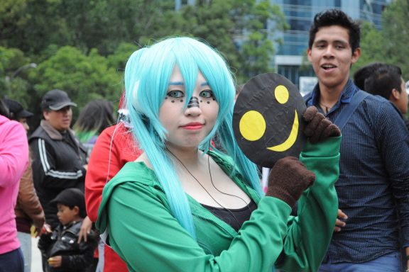 Person in blue wig and green hoodie pondering anime vs manga while participating in a cosplay event