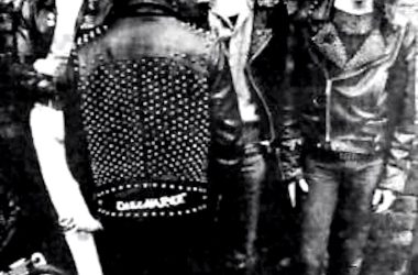 Black and white photo of four members of the punk band Discharge, with one member facing away, wearing a studded leather jacket with the band name on the back.