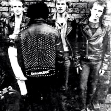 Black and white photo of four members of the punk band Discharge, with one member facing away, wearing a studded leather jacket with the band name on the back.