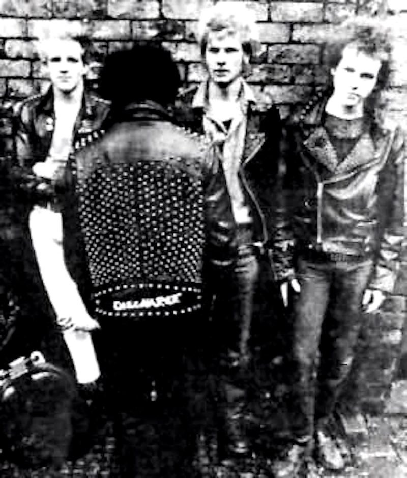 Black and white photo of four members of the punk band Discharge, with one member facing away, wearing a studded leather jacket with the band name on the back.