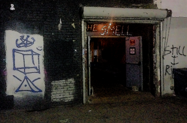 An entrance to a dimly lit los angeles punk venue building with punk clubs NYC graffiti on the surrounding walls and a sign above reading 'THE SMELL' in neon.